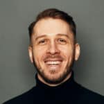 Studio photo of Alex, smiling and wearing a black turtleneck, on a gray background.