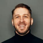 Studio photo of Alex, smiling and wearing a black turtleneck, on a gray background. 